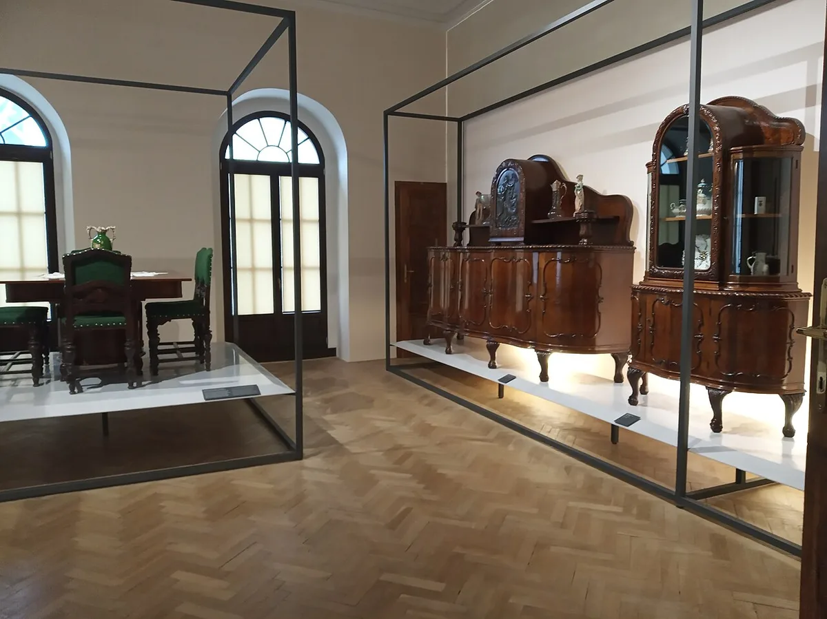 Een ingerichte museumkamer met een houten vitrinekast waarin blauw-wit porselein staat, naast een dressoir en een gedekte tafel met groene stoelen.