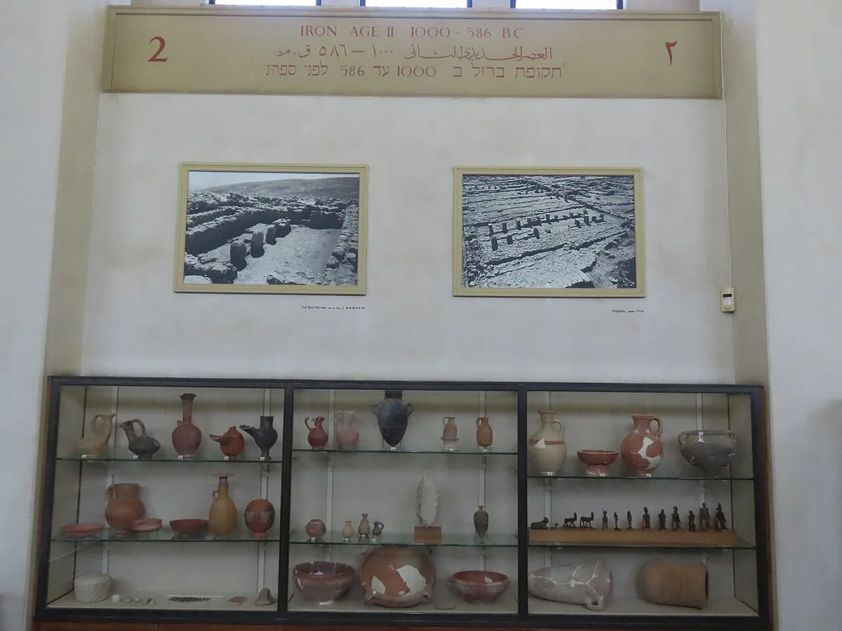 Een lange glazen wandvitrine in een museumzaal, gevuld met aardewerken kruiken en schalen op glazen legplanken; erboven hangen ingelijste foto's.