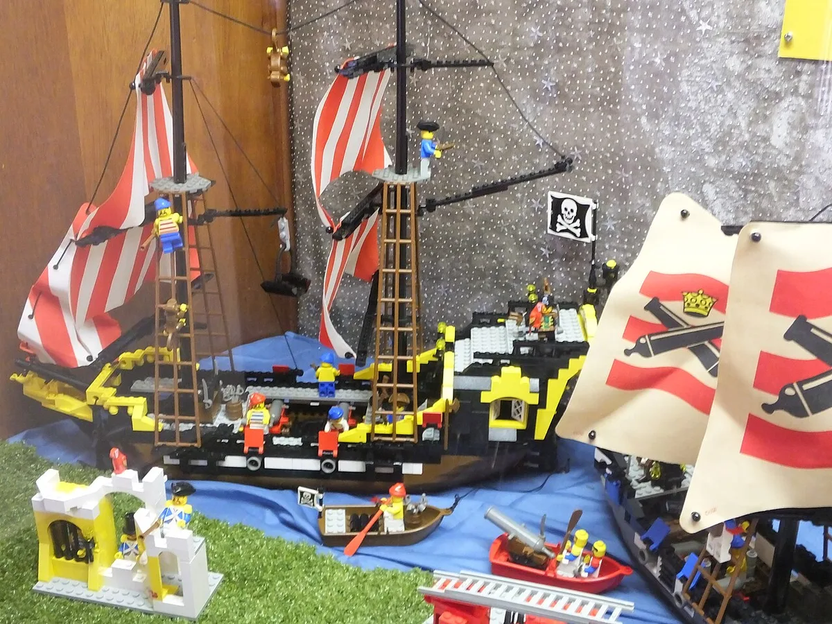 Een gebouwd LEGO-piratenschip met rood-wit gestreepte zeilen en minifiguren, opgesteld op een tafel naast andere LEGO-modellen.