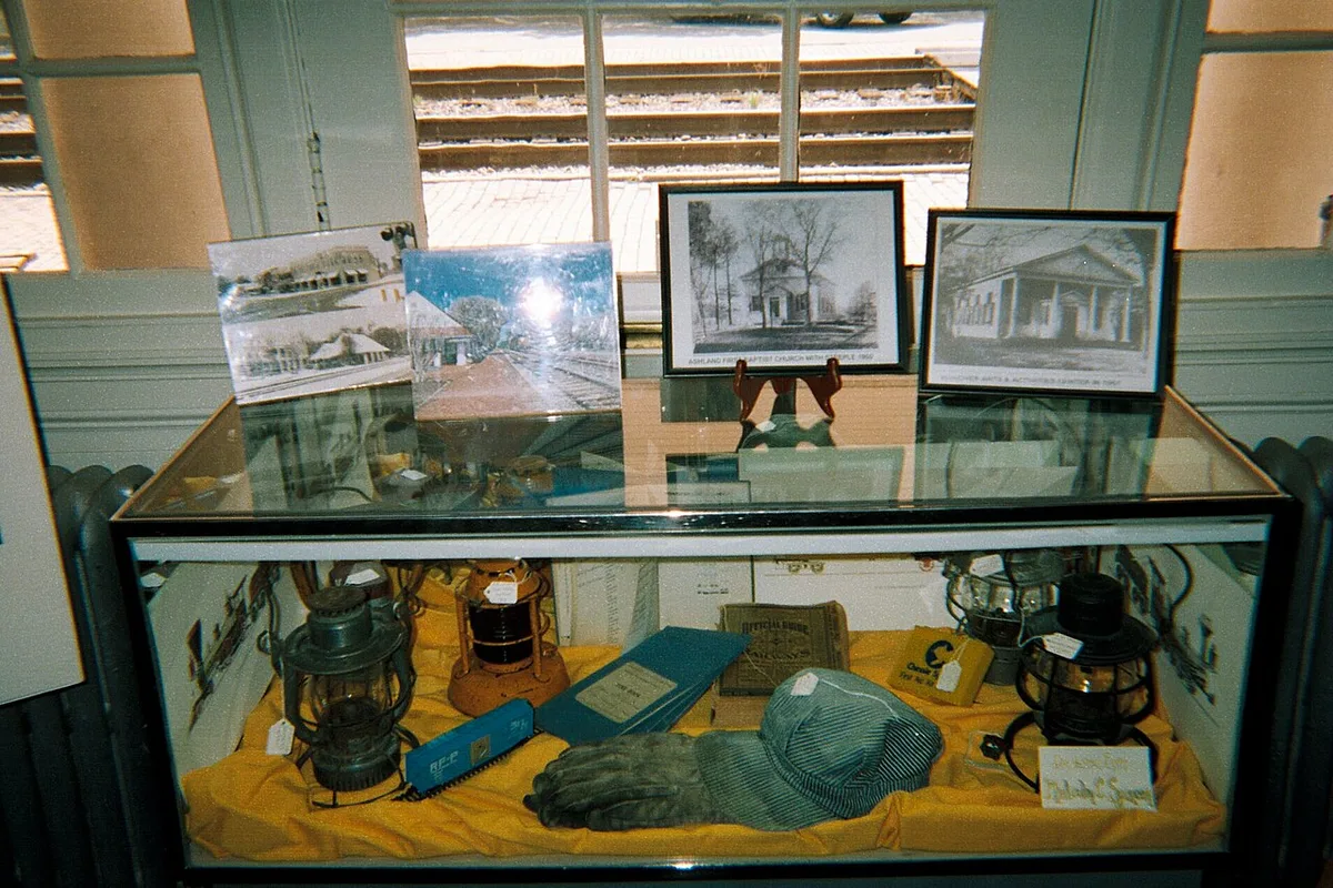 Een vitrine met glazen bovenblad vlak bij een zonnig raam; onder het glas liggen lantaarns, boeken en een pet, erbovenop staan ingelijste foto's.
