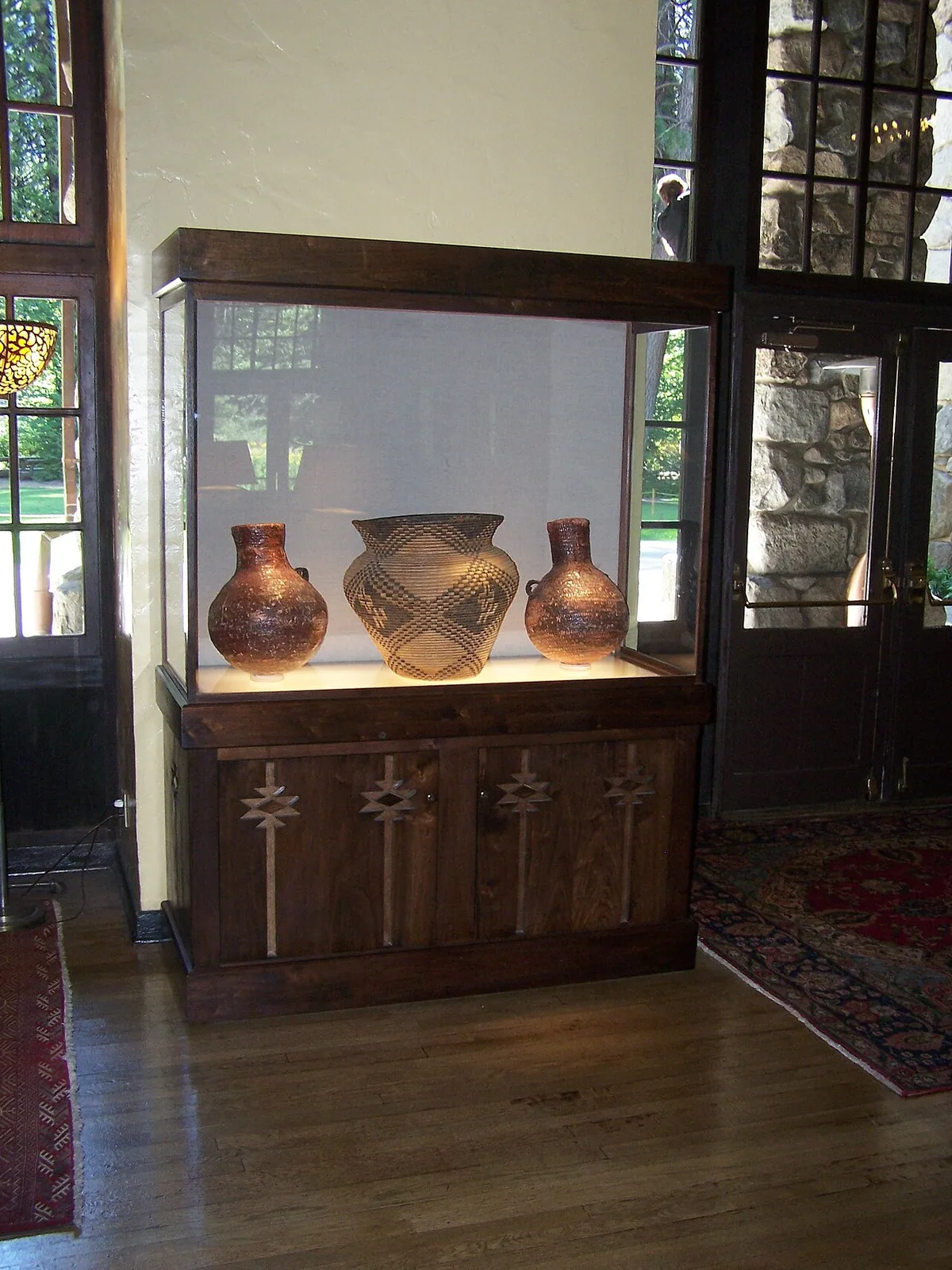 Een houten vitrinekast met glazen bovenbouw waarin drie beschilderde aardewerken potten staan; de kast staat op een gesloten onderkast in een woonkamer.